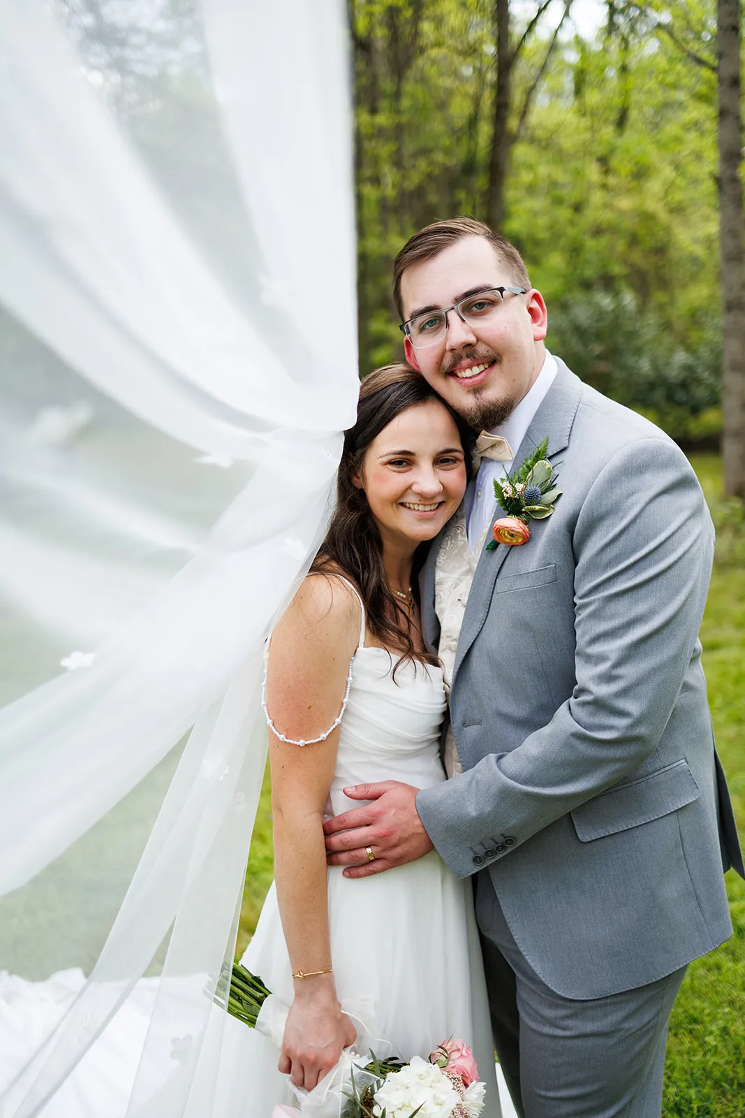 Couple smiling together beneath a flowing veil on the grounds at The Heritage Barn