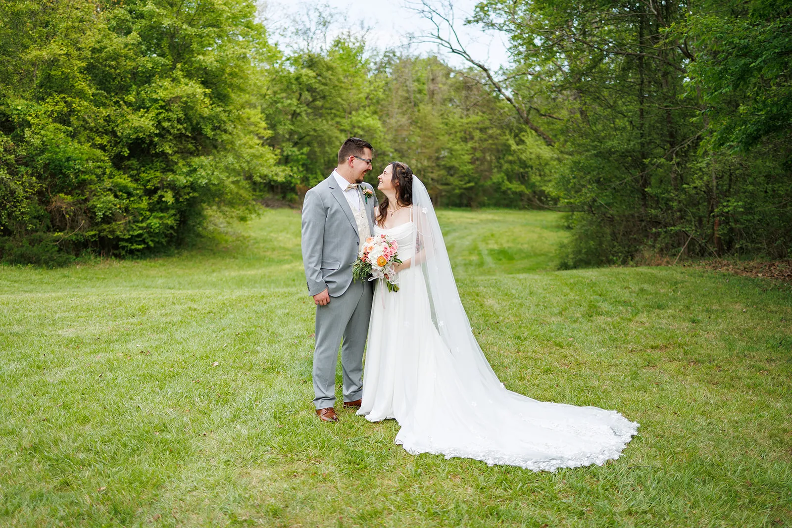 Couple standing together in a green field bordered by trees at The Heritage Barn